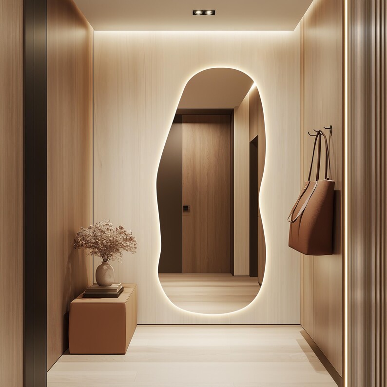 Puede incluir: Un pasillo moderno con paneles de madera clara y un gran espejo de forma org&aacute;nica con un borde brillante. Un bolso tote de cuero marr&oacute;n cuelga de la pared. Un peque&ntilde;o banco sostiene un jarr&oacute;n con flores secas.