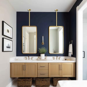 May include: A modern bathroom with a navy blue accent wall, featuring two rectangular gold-framed mirrors suspended from the ceiling. The vanity has light wood cabinets, a white countertop, and woven baskets below.
