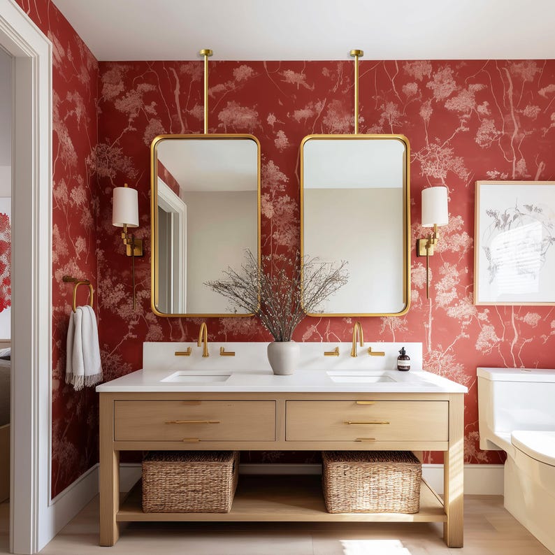 May include: A bathroom with a red floral wallpaper backdrop. Two rectangular gold-framed mirrors hang above a light wood vanity with a white countertop. Wicker baskets are below the vanity. Gold fixtures and sconces complete the design.