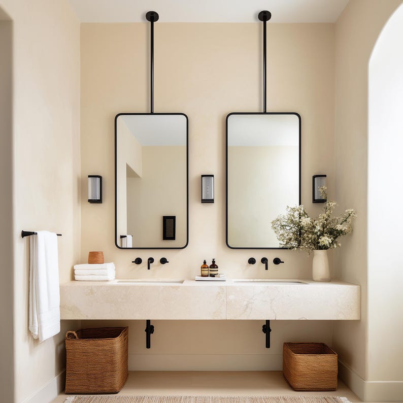 May include: A modern bathroom with two rectangular mirrors with black frames suspended from the ceiling. The mirrors are above a marble countertop with two sinks and black faucets. Wicker baskets and a vase of flowers add warmth.