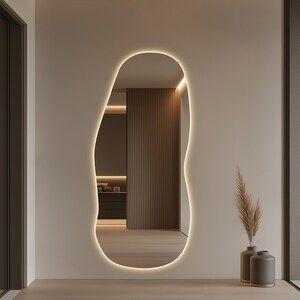 Puede incluir: Un espejo grande, de forma org&aacute;nica, con un borde LED brillante y c&aacute;lido, est&aacute; montado en una pared de tono neutro. El espejo refleja un interior moderno con detalles de madera e iluminaci&oacute;n suave. Dos jarrones grises con plantas secas se colocan en el suelo.