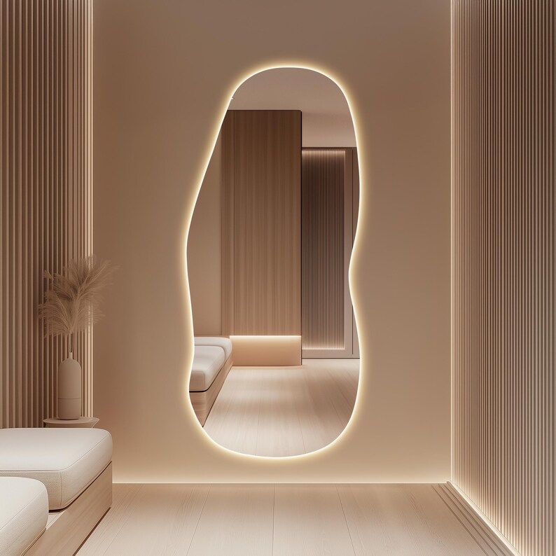 Puede incluir: Un espejo grande, de forma org&aacute;nica, con un borde brillante est&aacute; montado en una pared beige. El espejo refleja un interior minimalista con paneles y suelos de madera clara. La habitaci&oacute;n est&aacute; iluminada por una c&aacute;lida iluminaci&oacute;n empotrada.