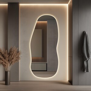 Puede incluir: Un espejo moderno de forma org&aacute;nica con un borde LED c&aacute;lido y brillante. El espejo est&aacute; colocado contra una pared de tonos neutros, complementado con un jarr&oacute;n oscuro con plantas secas y un suelo de madera. Una bata gris cuelga de una puerta.