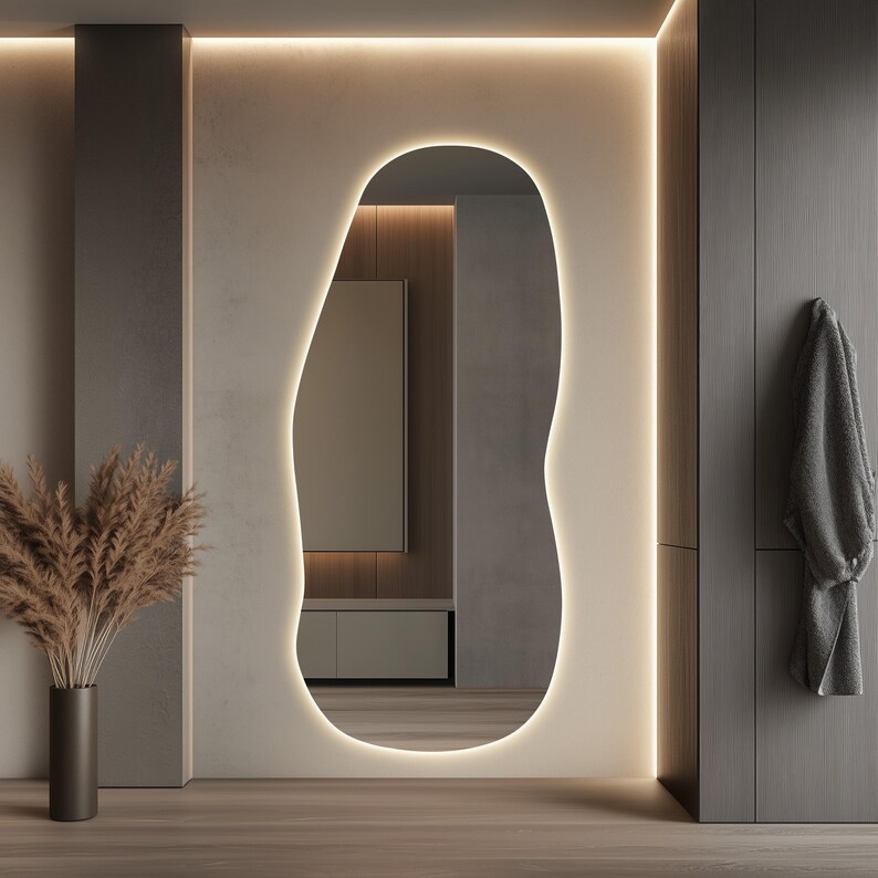 Puede incluir: Un espejo moderno de forma org&aacute;nica con un borde LED c&aacute;lido y brillante. El espejo est&aacute; colocado contra una pared de tonos neutros, complementado con un jarr&oacute;n oscuro con plantas secas y un suelo de madera. Una bata gris cuelga de una puerta.