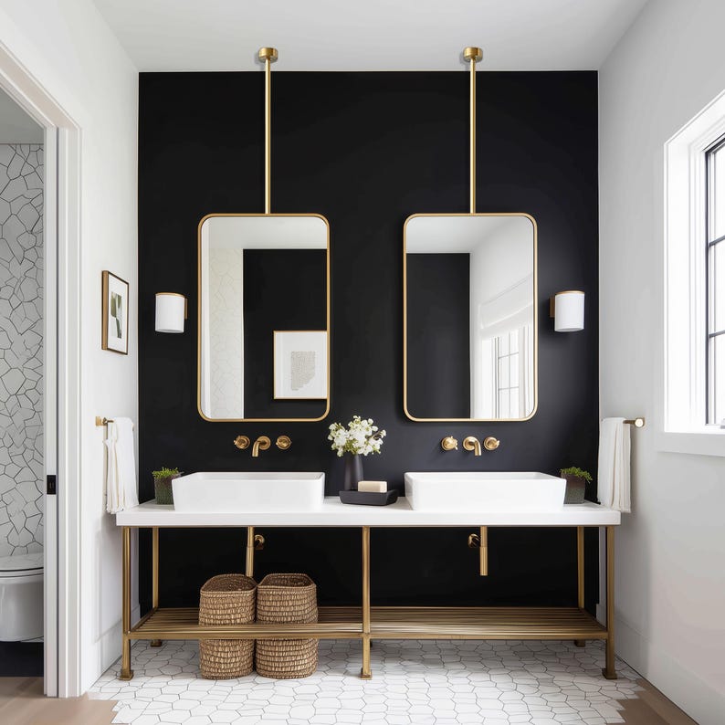 May include: A modern bathroom with a black accent wall features two rectangular gold-framed mirrors suspended from the ceiling. Two white sinks sit atop a gold-legged vanity. Woven baskets and a shelf are below the sinks. White towels hang on the side.