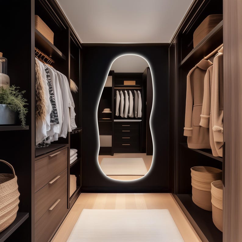 Puede incluir: Un vestidor con estanter&iacute;as y cajones de madera oscura. Un gran espejo de forma irregular con un borde blanco brillante est&aacute; centrado en la pared del fondo. La ropa cuelga de perchas y las cestas est&aacute;n en los estantes. El suelo es de madera clara.