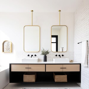 May include: A modern bathroom with two rectangular gold-framed mirrors suspended from the ceiling. The vanity features a black base, light wood drawers, and white sinks. Woven baskets are placed below the sinks, and a black vase holds greenery.