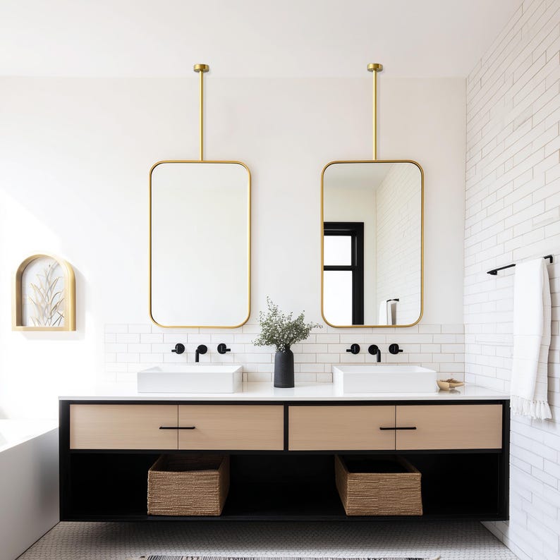 May include: A modern bathroom with two rectangular gold-framed mirrors suspended from the ceiling. The vanity features a black base, light wood drawers, and white sinks. Woven baskets are placed below the sinks, and a black vase holds greenery.