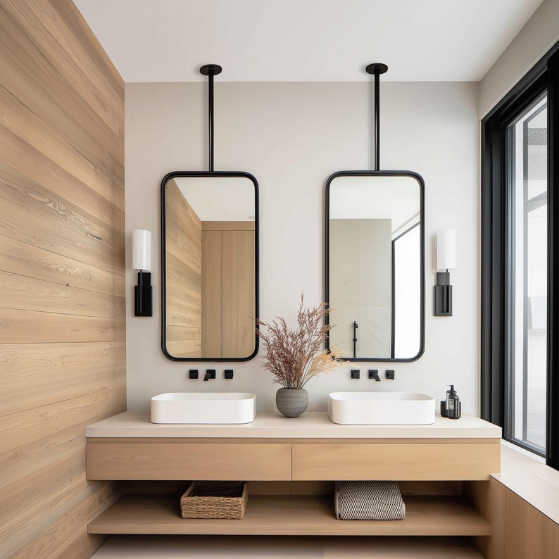 May include: A modern bathroom vanity with two rectangular mirrors suspended from the ceiling. The mirrors have black frames and are positioned above white rectangular sinks. The vanity is made of light wood, with a lower shelf and a basket.