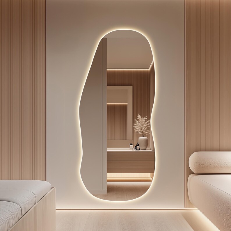 Puede incluir: Interior de dormitorio moderno con un gran espejo de forma org&aacute;nica con iluminaci&oacute;n integrada. El espejo refleja un tocador con un jarr&oacute;n y objetos decorativos. La habitaci&oacute;n tiene paneles de madera clara y una paleta de colores neutros.
