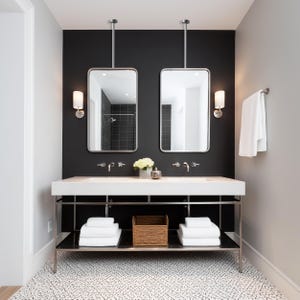 May include: A modern bathroom with a black accent wall, featuring two rectangular mirrors with silver frames suspended from the ceiling. A white countertop with two sinks, chrome faucets, and sconces. White towels and a woven basket are on the shelf below.