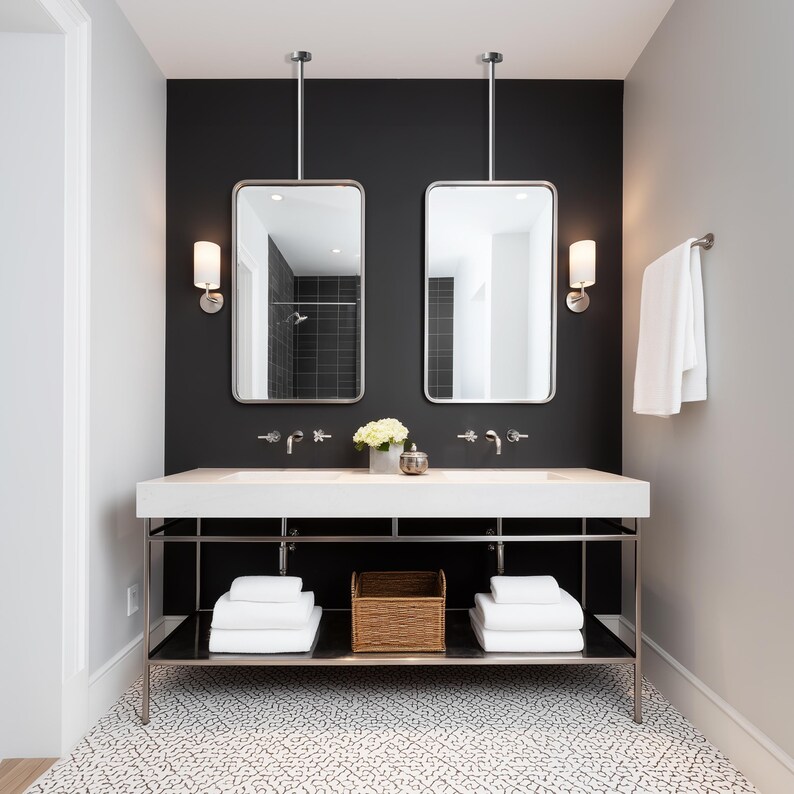 May include: A modern bathroom with a black accent wall, featuring two rectangular mirrors with silver frames suspended from the ceiling. A white countertop with two sinks, chrome faucets, and sconces. White towels and a woven basket are on the shelf below.
