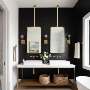 May include: A modern bathroom with a black accent wall, featuring two rectangular gold-framed mirrors suspended from the ceiling. The vanity has a white countertop, two white sinks, and a gold metal frame with a wooden shelf. Woven baskets sit below.