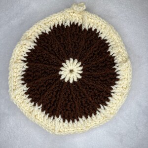 May include: A crocheted round cushion with a brown center and a white outer ring. The center has a white flower design.