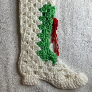 May include: White crocheted Christmas stocking with green and red trim. The stocking has a pointed toe and a green and red tassel.