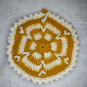 May include: A crocheted coaster in a yellow and white color scheme. The coaster has a six-pointed star design in the center and a scalloped edge.