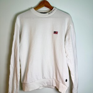 May include: A white sweatshirt with long sleeves, a crew neck, and a small American flag patch on the chest. The text "Marble Tree" is printed on the collar. The sweatshirt is hanging on a wooden hanger.