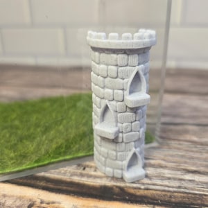 Op de afbeelding: Een kleine grijze kasteeltoren met een gekanteelde top en drie boogvensters. De toren is gemaakt van een steenachtig materiaal en is een decoratief item. De toren staat tegen een achtergrond van groen gras en een doorzichtige container.
