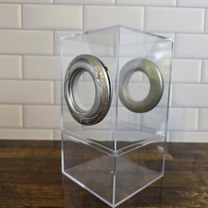 May include: Clear acrylic display case with two silver-toned, circular decorative elements. One element has an ornate floral design. The case sits on a dark wooden surface against a white tiled background.