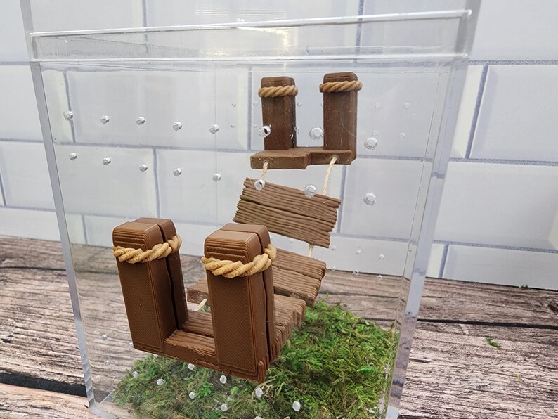 May include: A miniature wooden dock with a rope detail, set in a clear acrylic box with green moss and small white bubbles.