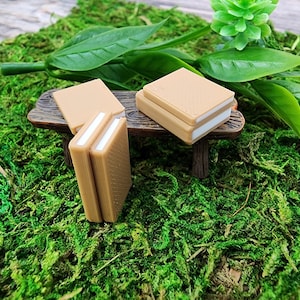 May include: Two stacks of miniature books on a small wooden bench. The books are brown with white pages and are arranged in a miniature garden setting.