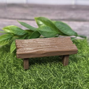 May include: A miniature brown wooden bench with a textured surface, resting on a bed of green artificial grass. Green leaves are placed behind the bench. The bench is a decorative item.
