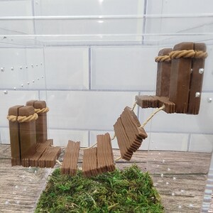 May include: A miniature wooden bridge with rope details, set in a clear plastic container with green moss at the base.