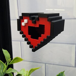 May include: A 3D printed heart-shaped object with a pixelated design. The heart is red with black outlines and has a small hole in the center. The heart is attached to a white tiled wall.
