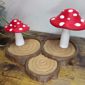 Può includere: Tre figurine decorative a forma di fungo. I funghi hanno cappelli rossi con pois bianchi e gambi bianchi. Sono inseriti in basi marroni a forma di ceppo d'albero. La composizione è su una superficie di legno.