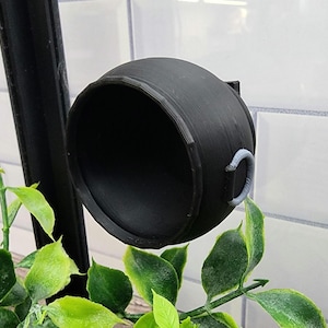 May include: A black cauldron with a grey handle, hanging from a black metal stand. The cauldron is in front of a white tiled wall.