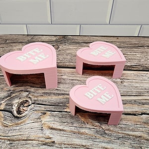 May include: Three pink heart-shaped stands with the words "Bite Me" printed in white on each. The stands are made of a smooth, glossy material and are perfect for displaying small items.