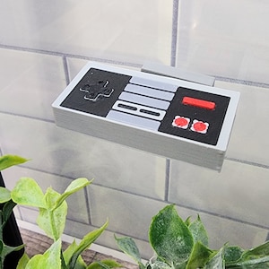 Puede incluir: Una réplica impresa en 3D de un mando de videojuego clásico, de color gris y negro. El mando tiene un pad direccional, seis botones y un botón de inicio rojo.
