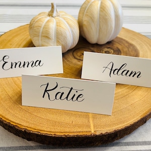 May include: Three white place cards with black calligraphy on a wooden slice. The place cards read "Emma", "Adam", and "Katie".