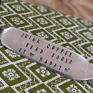 May include: A stamped metal bookmark with the words "DRINK COFFEE READ BOOKS BE HAPPY" in a black, all-caps font. The bookmark is on a green and white floral patterned background.