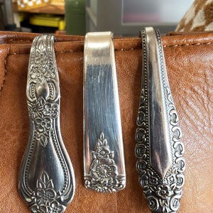 May include: Three silver spoon rests with ornate floral designs. The spoon rests are displayed on a brown leather surface. The spoon rests are made of silver metal and are in various shapes and sizes.