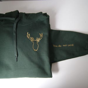 May include: A dark forest green hooded sweatshirt featuring a gold embroidered deer head outline on the chest and the text "You do not yield." embroidered in gold on the sleeve. The garment includes a matching drawstring hood and long sleeves.