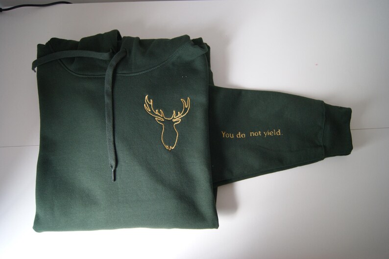May include: A dark forest green hooded sweatshirt featuring a gold embroidered deer head outline on the chest and the text "You do not yield." embroidered in gold on the sleeve. The garment includes a matching drawstring hood and long sleeves.