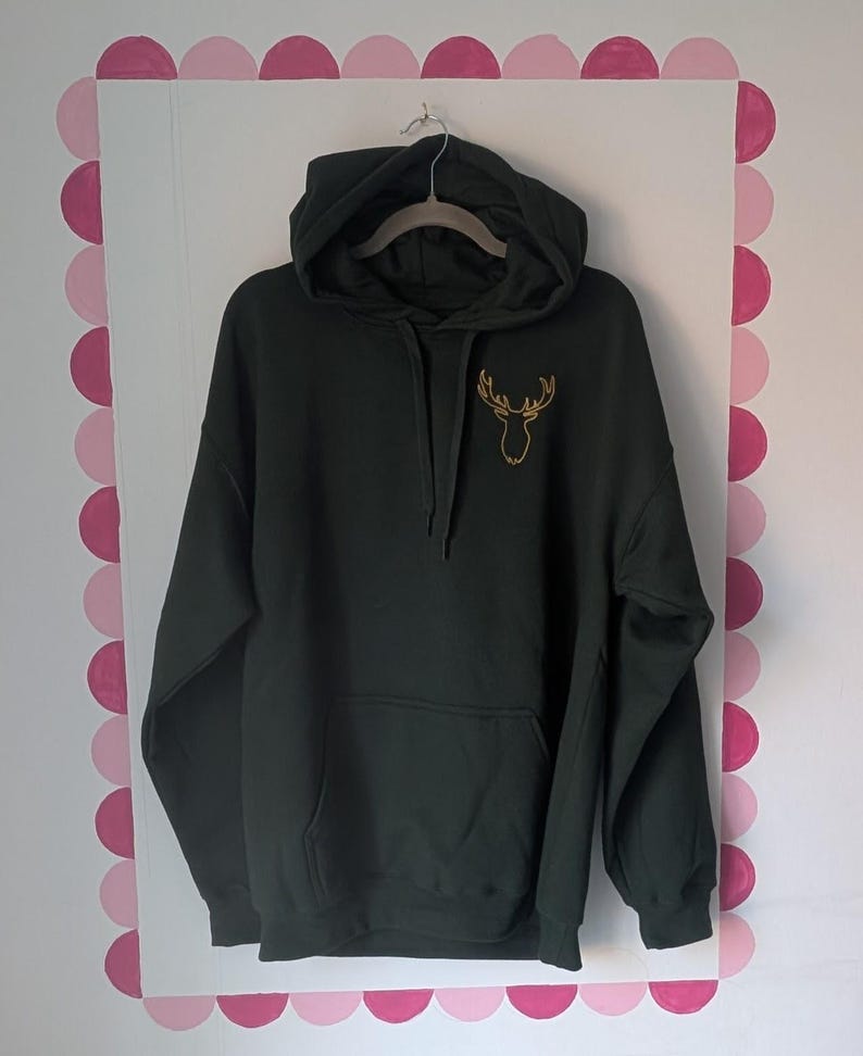 May include: A dark green hooded sweatshirt featuring a gold-colored embroidered outline of a deer head on the upper left chest. The garment includes a front kangaroo pocket, long sleeves with ribbed cuffs, and a matching drawstring at the hood.