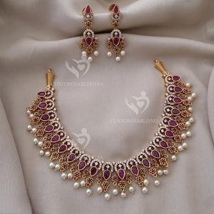 May include: A gold-coloured necklace and matching earrings set. The necklace and earrings feature teardrop-shaped, deep red gemstones, surrounded by small white stones and accented with small white pearls. The jewellery is displayed on a white fabric.