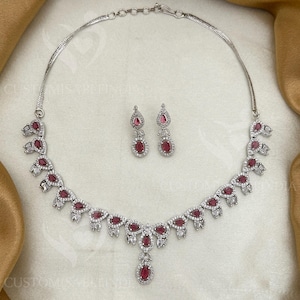 Può includere: Un set di collana e orecchini argentati. La collana presenta pietre rosse a forma di goccia e pietre di accento trasparenti. Gli orecchini coordinati hanno un design simile. Il set di gioielli è esposto su una superficie bianca.