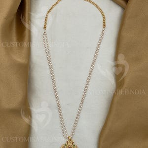 May include: A gold-colored necklace with a pendant featuring a central red gemstone, surrounded by smaller green and white stones. The pendant is suspended from a double strand of white pearls and a gold chain. The necklace is displayed on a beige fabric.