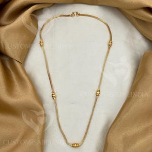 Puede incluir: Un collar dorado con un diseño de cadena fina y pequeñas cuentas redondas espaciadas uniformemente. El collar se muestra sobre un fondo de tela dorada.