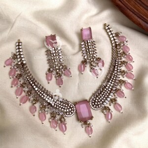 May include: A silver-toned necklace and matching earrings set. The necklace features a large, rectangular pink gemstone pendant and smaller pink beads. The earrings have a similar design with pink gemstones and dangling beads. The set is embellished with small, clear crystals.