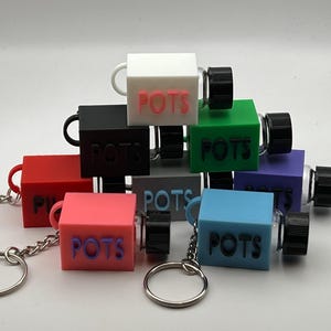 May include: A stack of eight colorful keychain containers with a small glass bottle attached to each. The containers are made of plastic and have the word "POTS" printed on them in various colors. Each container has a silver keychain attached.