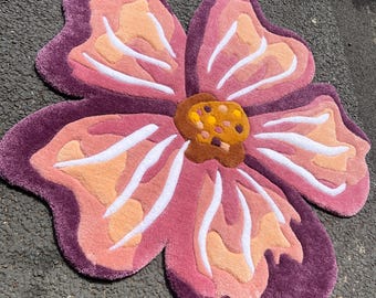 May include: A flower-shaped rug in shades of purple, pink, peach, and white. The rug's center is brown with yellow and orange details. The rug is made of a soft, plush material. The rug's design is layered, creating a 3D effect.