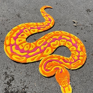 Könnte beinhalten: Ein getufteter Teppich in Schlangenform mit einem lebendigen Muster aus leuchtend orangefarbenen, gelben und pinkfarbenen Flecken. Die Schlange ist in einer geschwungenen, gewundenen Haltung dargestellt, mit strukturierter Oberfläche und Kopf.