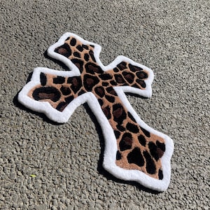 Puede incluir: Una alfombra de estampado de leopardo marrón y negro con forma de cruz y borde blanco. La alfombra está sobre una superficie de asfalto gris.
