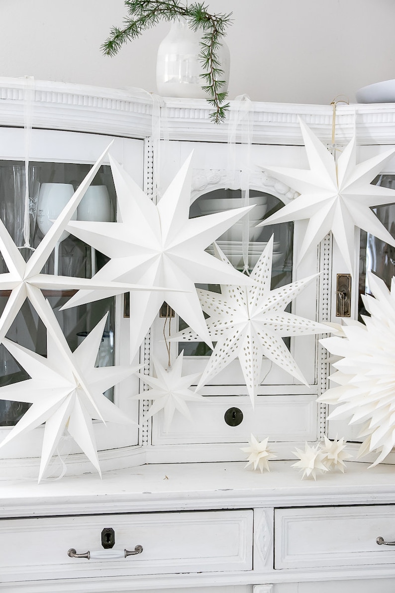 May include: A collection of white paper star ornaments in various sizes, featuring both solid and perforated designs. These multi-pointed stars are suspended by thin ribbons, creating a layered display of geometric shapes with sharp, clean edges.