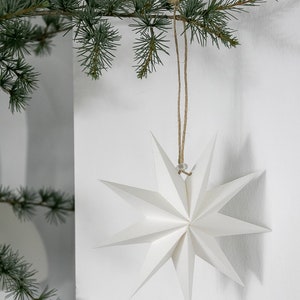 May include: A white, multi-pointed paper star ornament with sharp, geometric edges. The star is suspended by a thin, natural-colored jute twine string, which is looped over a green pine branch, creating a clean and simple decorative display.