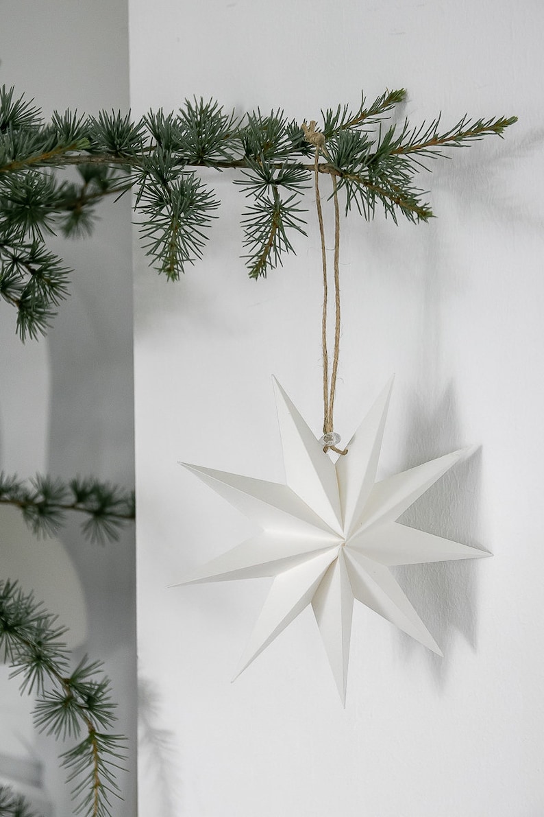 May include: A white, multi-pointed paper star ornament with sharp, geometric edges. The star is suspended by a thin, natural-colored jute twine string, which is looped over a green pine branch, creating a clean and simple decorative display.